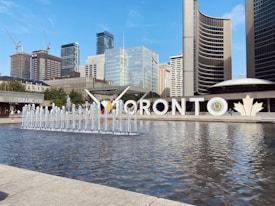 A large sign spelling 'Toronto' stands prominently in front of a reflecting pool with multiple water fountains. The surrounding area features modern skyscrapers and the uniquely curved structure of the City Hall. A clear blue sky provides a bright and welcoming backdrop. A snack pavilion is visible on the left.