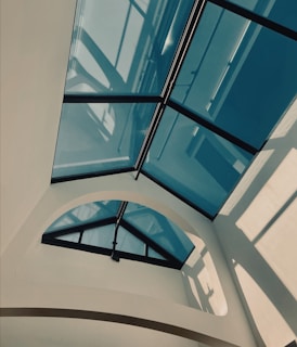 A modern architectural design featuring a glass ceiling with triangular panes and dark metal frames. Soft natural light filters through, creating geometric shadows on the cream-colored walls.