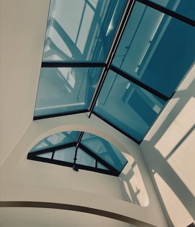 A modern architectural design featuring a glass ceiling with triangular panes and dark metal frames. Soft natural light filters through, creating geometric shadows on the cream-colored walls.