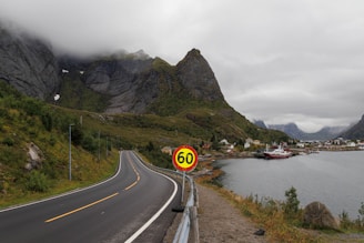 A winding road curves along a fjord, with a speed limit sign displaying 60. Towering mountains clad in mist create a dramatic backdrop, while a small village with red and white houses nestles by the water. A large boat is docked near the shoreline, enhancing the scenic coastal atmosphere.
