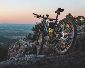 A mountain bike is perched on a rocky outcrop overlooking a wide, picturesque natural landscape during sunset. The scene includes rugged terrain with some greenery and a distant view of mountains under a clear sky transitioning from pale blue to soft pink hues.