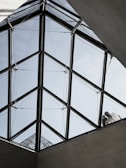 A geometric, triangular glass roof structure with clear panels, supported by a metal framework. Two figures are visible standing on the structure, partially obscured by the glass, interacting with the roof. The background shows a pale blue sky.