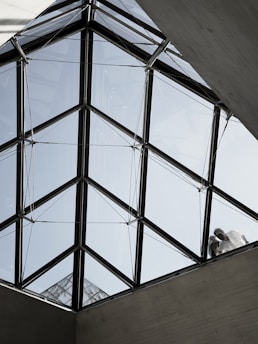A geometric, triangular glass roof structure with clear panels, supported by a metal framework. Two figures are visible standing on the structure, partially obscured by the glass, interacting with the roof. The background shows a pale blue sky.