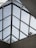 A geometric, triangular glass roof structure with clear panels, supported by a metal framework. Two figures are visible standing on the structure, partially obscured by the glass, interacting with the roof. The background shows a pale blue sky.