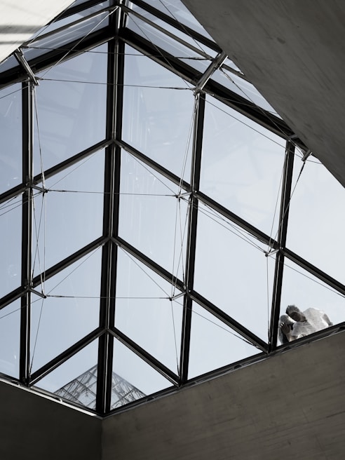 A geometric, triangular glass roof structure with clear panels, supported by a metal framework. Two figures are visible standing on the structure, partially obscured by the glass, interacting with the roof. The background shows a pale blue sky.