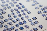 Numerous scattered puzzle pieces lie on a flat, light-colored surface. The pieces are predominantly blue with a glossy finish, appearing randomly distributed across the surface.