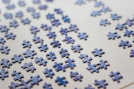 Numerous scattered puzzle pieces lie on a flat, light-colored surface. The pieces are predominantly blue with a glossy finish, appearing randomly distributed across the surface.