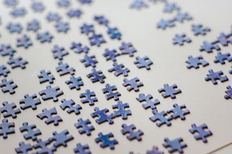 Numerous scattered puzzle pieces lie on a flat, light-colored surface. The pieces are predominantly blue with a glossy finish, appearing randomly distributed across the surface.