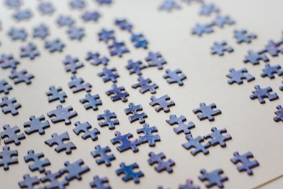 Numerous scattered puzzle pieces lie on a flat, light-colored surface. The pieces are predominantly blue with a glossy finish, appearing randomly distributed across the surface.