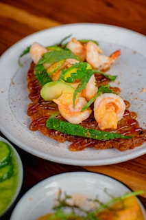 A gourmet dish featuring shrimp on a bed of thinly sliced avocado and herbs, garnished with fine greenery, and served on a white ceramic plate. The background shows hints of another dish with green sauce, also presented on a dish.