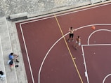 An aerial view of a basketball court with two shirtless individuals playing basketball. A basketball is in motion, with one player attempting a shot. Another person, sitting on stone steps nearby, appears to be spectating or taking a break. Various personal items, including a bag and shoes, are placed on the steps.