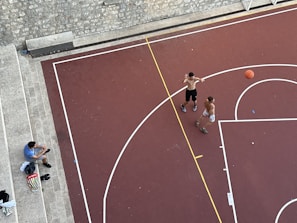 An aerial view of a basketball court with two shirtless individuals playing basketball. A basketball is in motion, with one player attempting a shot. Another person, sitting on stone steps nearby, appears to be spectating or taking a break. Various personal items, including a bag and shoes, are placed on the steps.