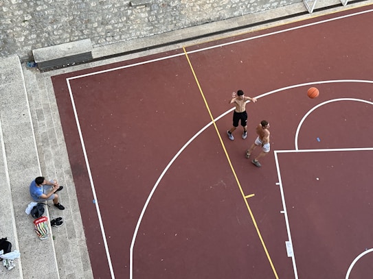 An aerial view of a basketball court with two shirtless individuals playing basketball. A basketball is in motion, with one player attempting a shot. Another person, sitting on stone steps nearby, appears to be spectating or taking a break. Various personal items, including a bag and shoes, are placed on the steps.