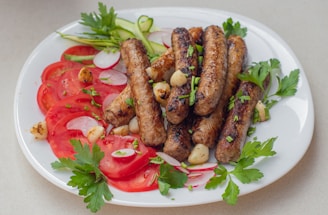 A white plate filled with grilled sausages, tomato slices, radish slices, parsley, garlic cloves, and cucumber ribbons arranged in an appetizing manner. The sausages are garnished with chopped herbs, and the vibrant colors of the vegetables add freshness to the dish.