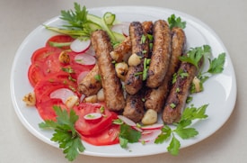 A white plate filled with grilled sausages, tomato slices, radish slices, parsley, garlic cloves, and cucumber ribbons arranged in an appetizing manner. The sausages are garnished with chopped herbs, and the vibrant colors of the vegetables add freshness to the dish.