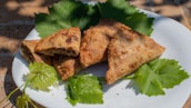 Three baked triangular pastries are placed on a white plate, surrounded by green grape leaves. The pastries have a golden-brown, crispy exterior and appear to be filled with a savory or sweet mixture.