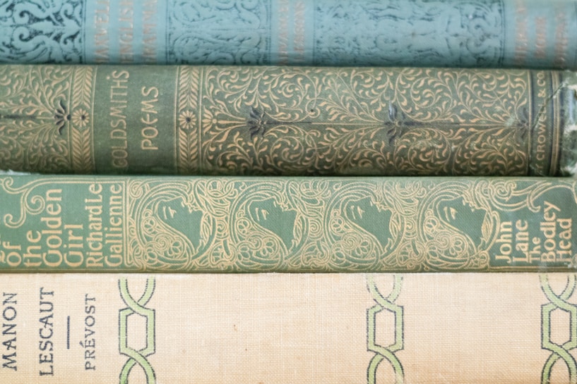A stack of vintage book spines, each with distinct ornate designs and faded embossing. The colors are primarily shades of green and beige, with intricate patterns featuring floral and human figure motifs. The texts on the spines are elegantly styled, suggesting classic literary works.