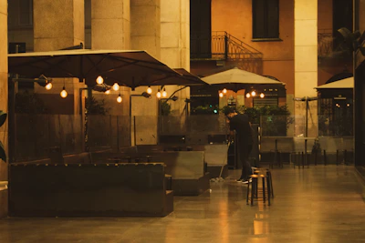 A dimly lit outdoor seating area with multiple large umbrellas and glowing hanging light bulbs. There are plants and glass partitions along the perimeter. A person is seen cleaning the floor, wearing dark clothing. The setting appears serene and quiet, with reflections visible on the polished floor.