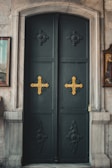A large, ornate dark green door with two prominent golden crosses, set in a stone doorway. The door features intricate carved designs around the crosses and has a marble frame with textured stone walls on either side. There are framed pictures visible partially on the left and right walls adjoining the door.