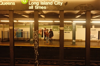 An underground subway platform with signage indicating trains to Long Island City. Several people are standing on the platform, including two individuals near the center. The station has industrial lighting with tiled walls displaying advertisements.