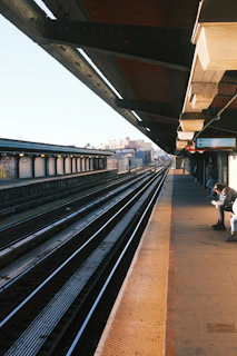 An elevated train station platform with multiple railway tracks running parallel. The platform stretches into the distance with a canopy providing partial shade. A person sits on the platform bench, possibly looking at a device.