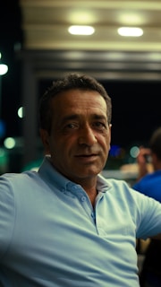 A middle-aged man wearing a light blue polo shirt sits in a dimly lit setting, likely at an outdoor cafe or similar venue at night. The background is blurred with bokeh lights, suggesting an urban environment with some people visible.