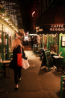 A narrow street is lined with small cafes and restaurants, illuminated by warm, glowing string lights. A person with long hair is walking away, carrying two large bags in their hands. The signage of a cafe reading 'Caffe Reggio' is prominently visible, alongside chalkboard menus and small, outdoor seating areas.