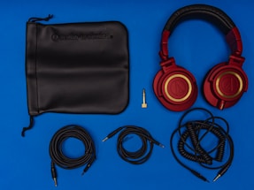 A set of red and gold over-ear headphones with a black leather carrying pouch lies beside various audio cables and a gold-plated audio adapter on a blue background. The cables include a coiled cable, a straight cable, and an additional cable with connectors.