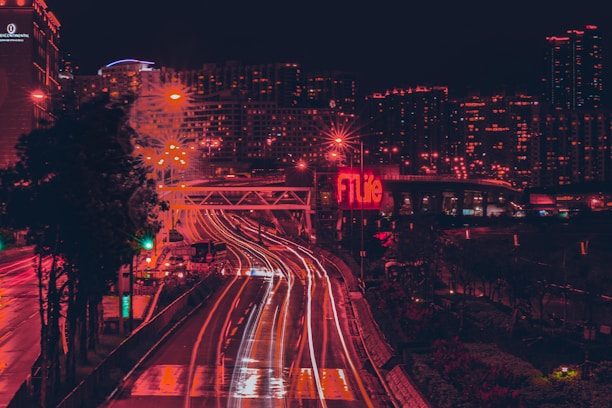 A nighttime cityscape featuring a long-exposure shot of a busy highway with streaks of car lights creating dynamic lines. Buildings in the background are illuminated with numerous windows visible. A prominent neon sign with the word 'FTLife' is visible, casting a red hue over the scene.