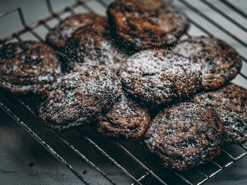 A batch of freshly baked cookies is cooling on a metal rack, sprinkled with a generous dusting of powdered sugar. The cookies have a rich, dark color, suggesting they might be chocolate-flavored or similar.