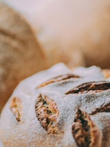 Close-up of freshly baked artisan bread with a golden-brown crust and a dusting of flour. The bread features deep cuts or slashes on the surface, highlighting its rustic texture.