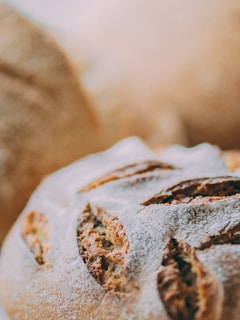 Close-up of freshly baked artisan bread with a golden-brown crust and a dusting of flour. The bread features deep cuts or slashes on the surface, highlighting its rustic texture.