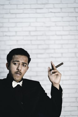 A person wearing a bow tie and dark suit holds a cigar in their hand against a white brick wall backdrop. Their expression appears confident and contemplative.