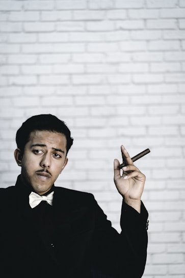 A person wearing a bow tie and dark suit holds a cigar in their hand against a white brick wall backdrop. Their expression appears confident and contemplative.