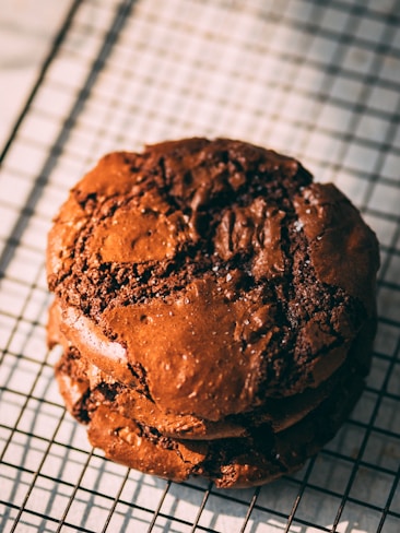 A stack of rich, chocolate cookies with a cracked surface, resting on a cooling rack. The texture appears chewy and moist, suggesting freshly baked goodness. Light shines on the cookies, highlighting their glossy and slightly uneven surface.