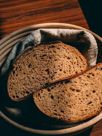 Two slices of rustic brown bread are placed on a cloth inside a round wooden basket. The bread has a porous texture, indicating it may be sourdough or a similar type. The warm lighting casts gentle shadows, enhancing the color of the bread and the wooden table surface beneath.