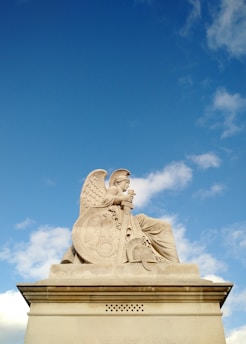 A marble statue of a seated angel with large wings and a serene expression. The angel is holding a shield and a long staff or spear. The carving details include intricate designs on the shield and the angel's robe. The statue is set against a backdrop of a bright blue sky with scattered clouds.