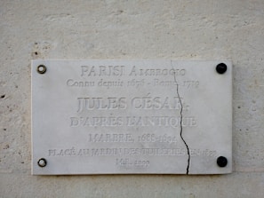 A marble plaque mounted on a wall features engraved text related to Julius Caesar. The plaque is cracked vertically near the right side and is secured with visible screws at each corner. The background is a textured stone surface.
