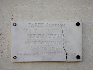 A marble plaque mounted on a wall features engraved text related to Julius Caesar. The plaque is cracked vertically near the right side and is secured with visible screws at each corner. The background is a textured stone surface.