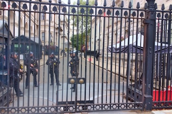 Five uniformed officers are standing behind a tall, ornate metal gate. The setting appears to be an official or governmental building with grand architecture, including columns and windows with flower boxes. One officer appears to be standing at the gate while the others are gathered in a group further back. In the background, there are security booths and a large canopy structure.