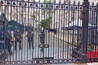 Five uniformed officers are standing behind a tall, ornate metal gate. The setting appears to be an official or governmental building with grand architecture, including columns and windows with flower boxes. One officer appears to be standing at the gate while the others are gathered in a group further back. In the background, there are security booths and a large canopy structure.
