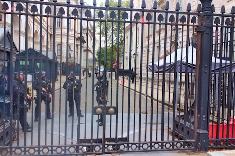 Five uniformed officers are standing behind a tall, ornate metal gate. The setting appears to be an official or governmental building with grand architecture, including columns and windows with flower boxes. One officer appears to be standing at the gate while the others are gathered in a group further back. In the background, there are security booths and a large canopy structure.