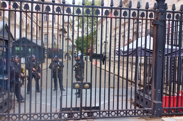 Five uniformed officers are standing behind a tall, ornate metal gate. The setting appears to be an official or governmental building with grand architecture, including columns and windows with flower boxes. One officer appears to be standing at the gate while the others are gathered in a group further back. In the background, there are security booths and a large canopy structure.