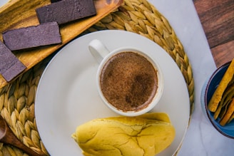 A cup of frothy hot chocolate rests on a white plate accompanied by a soft bread roll. To the side, dark chocolate bars are arranged on a wooden tray, and crispy breadsticks are placed in a small blue bowl. The setting includes a woven mat and wooden accents, creating a cozy and inviting atmosphere.
