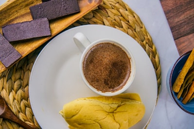 A cup of frothy hot chocolate rests on a white plate accompanied by a soft bread roll. To the side, dark chocolate bars are arranged on a wooden tray, and crispy breadsticks are placed in a small blue bowl. The setting includes a woven mat and wooden accents, creating a cozy and inviting atmosphere.