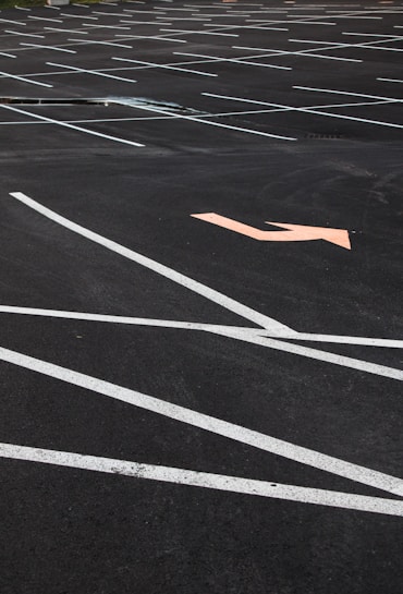An empty parking lot with neatly painted white lines indicating parking spaces. An orange arrow on the asphalt points in a direction, offering guidance for traffic flow. The surface is smooth with no visible vehicles or people present.