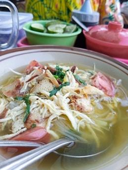 A close-up view of a bowl of soup containing shredded chicken, noodles, and garnished with fresh herbs. The soup is served in a ceramic bowl with visible green garnishes and pieces of meat. In the background, there are containers of sliced limes and vibrant, colorful cloths.