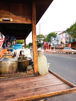 A wooden street food cart is placed by the roadside, featuring metal kettles and a plastic container. Beyond the cart, a road with passing cars is visible, flanked by trees and storefronts. The area looks active and urban, highlighted by clear skies and some banners or flags on the buildings.