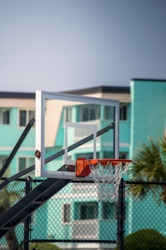 A basketball hoop with a transparent backboard is set against the backdrop of a teal-colored building and a clear sky. The hoop and net are attached to a black metal frame, and a chain-link fence is visible in the foreground.