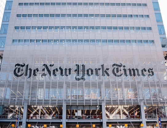 The building facade features large, modern architectural elements with a grid-like structure. The name of a well-known newspaper is prominently displayed in bold, black letters across the midsection. The structure consists of multiple floors with visible windows, suggesting it is an office building. The design incorporates a sleek and contemporary style.
